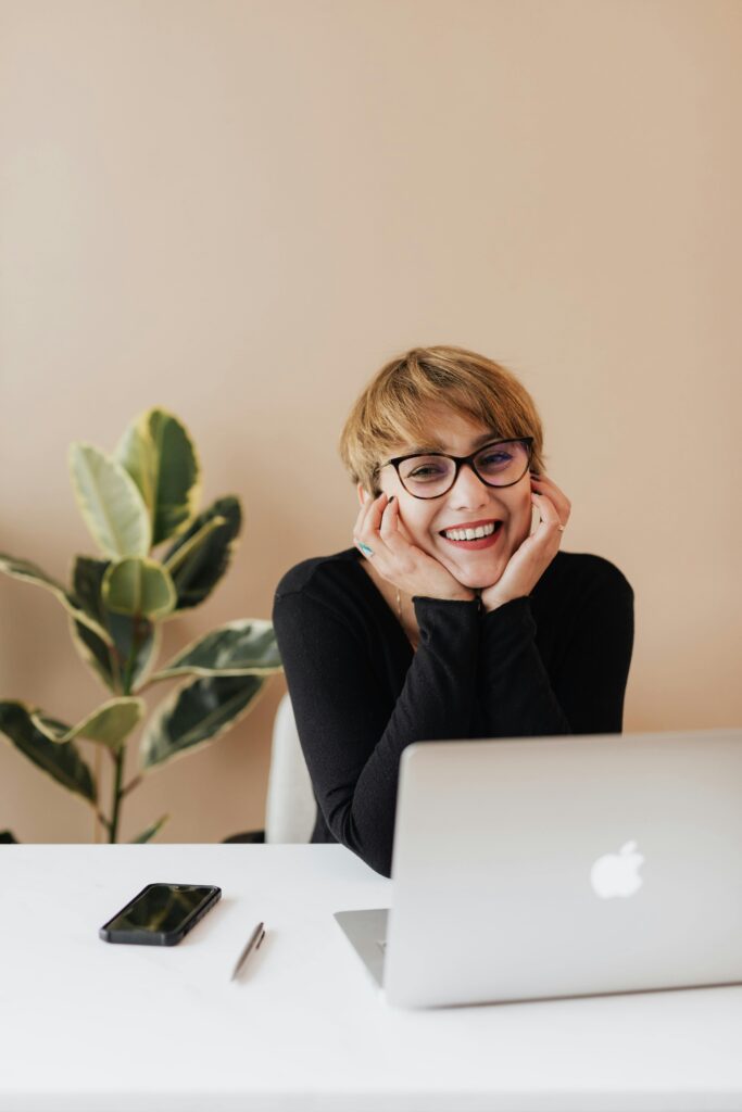 Cheerful woman in eyeglasses and black sweater smiling while working at a desk with a laptop indoors.